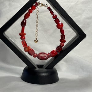 May include: A red beaded bracelet with a gold-colored clasp and extender chain. The bracelet features various shapes and sizes of red beads, displayed in a black, diamond-shaped frame.