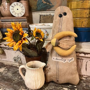 May include: A tan burlap ghost figurine with two black button eyes, a yellow fabric crescent moon accent, and a small label reading a haunting night. Beside it sits a small cream-colored ceramic pitcher and a potted sunflower arrangement.