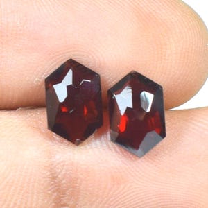 May include: Two loose, deep red, hexagonal-cut gemstones with faceted surfaces. The stones exhibit a dark, rich crimson hue and a polished finish, reflecting light across their geometric, multi-sided shapes, showcasing clarity and depth.