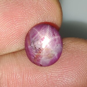 May include: A round, cabochon-cut gemstone with a deep magenta and pink hue. The stone features a visible star-like optical phenomenon known as asterism, with light reflecting across its polished, translucent surface in a distinct pattern.