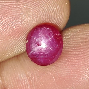Puede incluir: Una piedra preciosa redonda tallada en cabujón de un color rojo magenta profundo y vibrante. La superficie de la piedra muestra una textura interna fibrosa sutil y un reflejo de luz centrado y brillante, lo que indica un acabado liso, pulido y abombado.