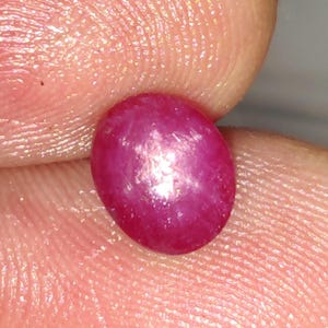 Puede incluir: Una gema de rubí ovalada y vibrante. La superficie pulida refleja la luz, mostrando un tono rojo intenso. La gema se sostiene entre dos dedos, resaltando su tamaño y forma. Ideal para la creación de joyas.