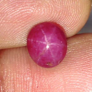 Puede incluir: Primer plano de una vibrante gema de rubí de forma ovalada. La gema es de un color rojo rosado intenso, con un patrón de estrella de seis rayos visible en su superficie. El rubí se sostiene entre dos dedos, mostrando su calidad pulida y reflectante.