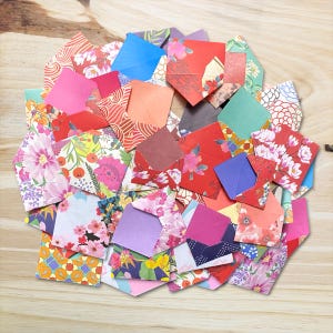 May include: A pile of colourful origami envelopes with floral and patterned designs. The envelopes are in shades of pink, red, orange, blue, and green, arranged on a light wood surface.