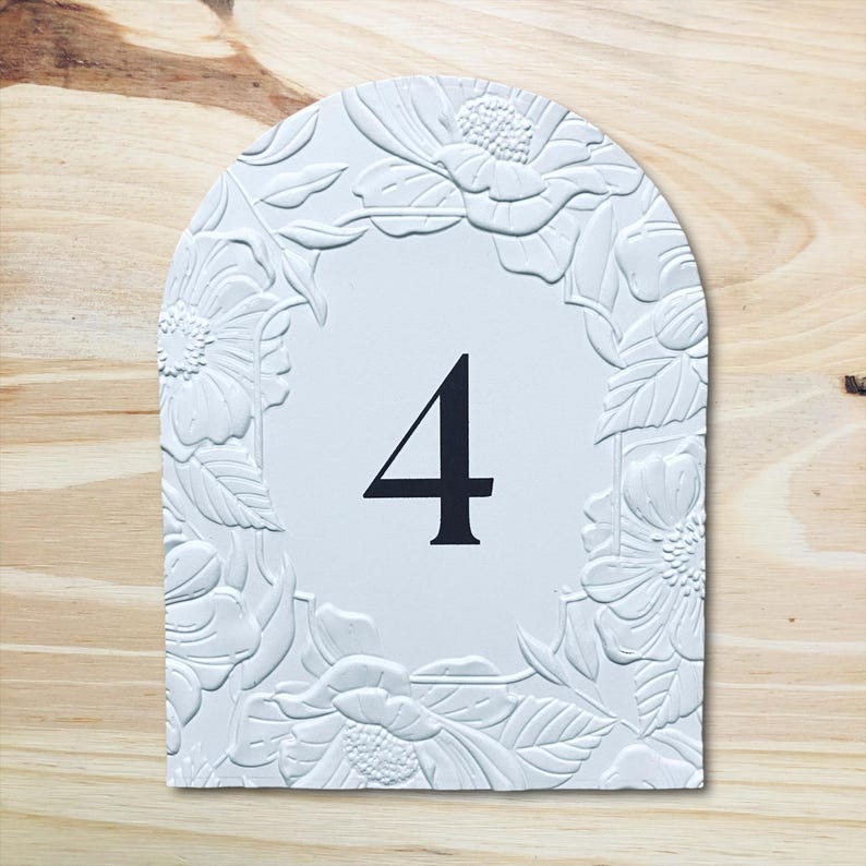 May include: White, arched table number four with a floral embossed border. The number is in black, centered on a white background. The background is a light wood grain.