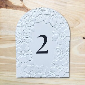 May include: White, arched table number 2, framed by embossed floral designs. The number is in black, centered on the white surface. The background is a light wood grain.