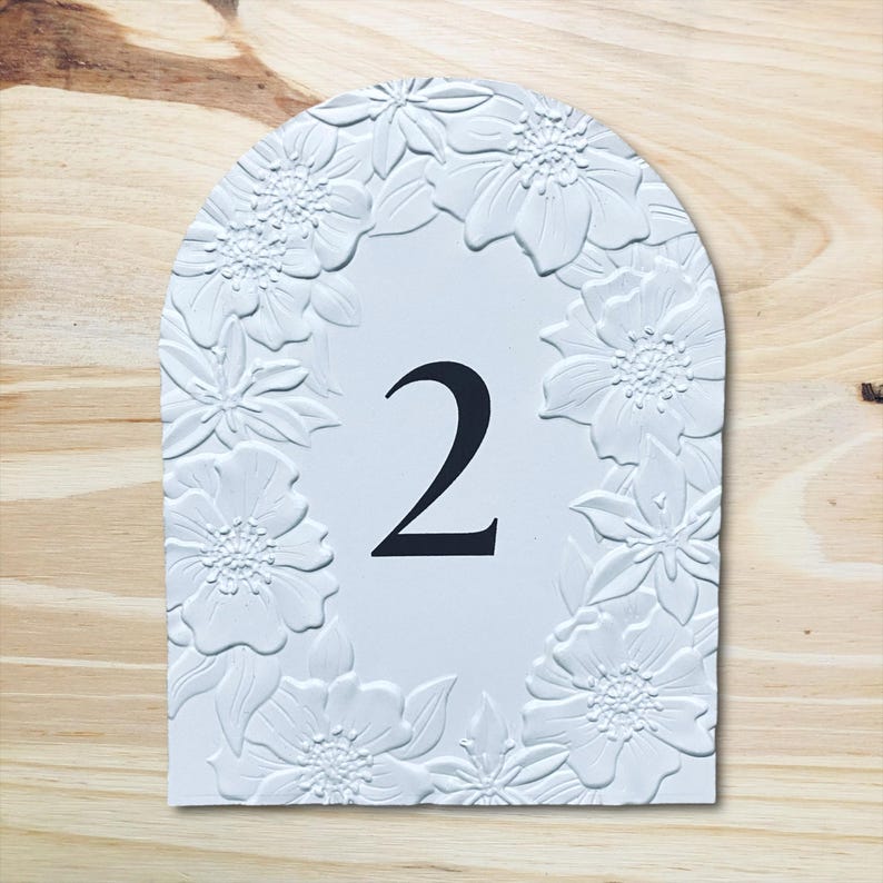May include: White, arched table number 2, framed by embossed floral designs. The number is in black, centered on the white surface. The background is a light wood grain.
