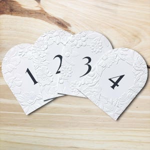 May include: Four white heart-shaped table number cards with embossed floral designs and black numerals 1, 2, 3, and 4. The cards are arranged on a light wood surface, suitable for weddings or events.