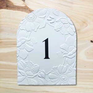 May include: White, arched sign with a floral border and the number 1 in black. The flowers are embossed, creating a textured effect. The sign is set against a light wood grain background.