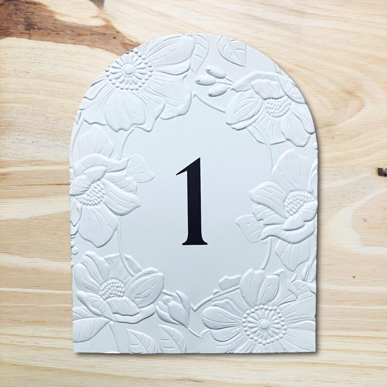 May include: White, arched sign with a floral border and the number 1 in black. The flowers are embossed, creating a textured effect. The sign is set against a light wood grain background.