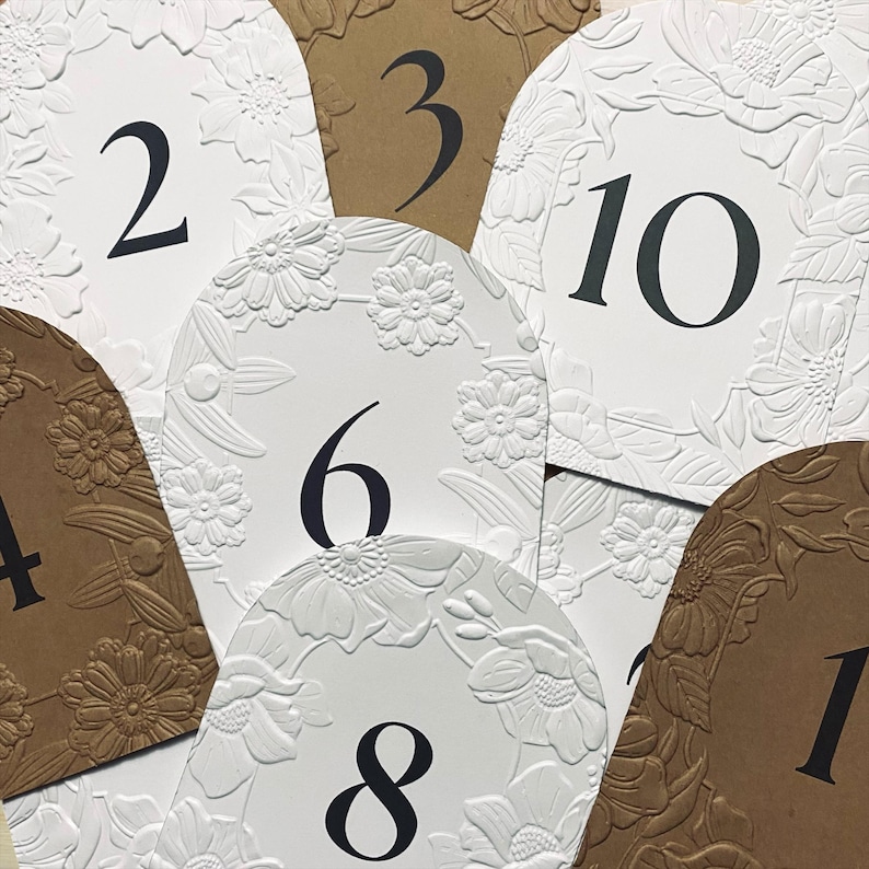 May include: Close-up of several table number cards. The cards are in various shapes and colors, including white and brown. Each card features a large, black number and a floral embossed design. The numbers range from 1 to 10.