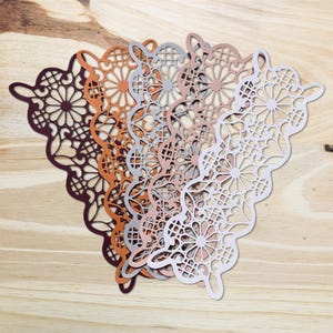 May include: A collection of intricately cut paper doilies in various colors, including deep burgundy, burnt orange, silver, rose gold, and white. The doilies feature a detailed floral and geometric pattern, arranged on a light wood surface.