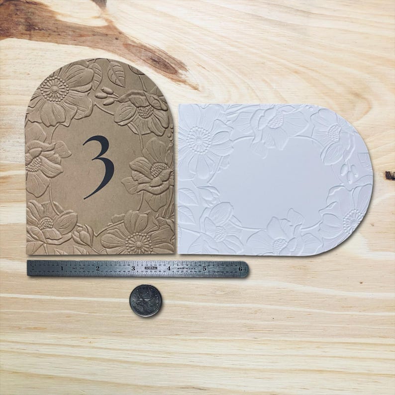 May include: Two table numbers with floral designs. One is tan with the number 3 in black, the other is white. A metal ruler shows measurements in inches. A quarter is also present.