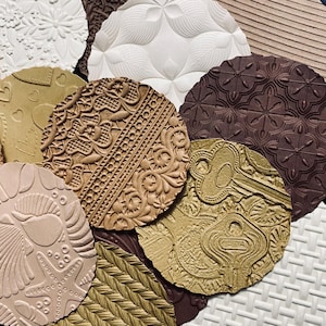 May include: A selection of circular, textured craft supplies in various colours. Designs include floral patterns, keys, hearts, and geometric shapes. Colours range from white and tan to brown and dark burgundy.