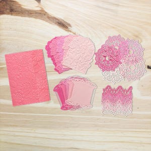 May include: A selection of pink and light pink paper craft items, including embossed floral cards, die-cut lace doilies, and decorative shapes. The items are arranged on a light wood grain surface.