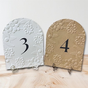 May include: Two decorative table numbers, featuring a floral design. One is white with the number 3 in black, the other is tan with the number 4 in black. Both have a curved top and decorative metal stands.