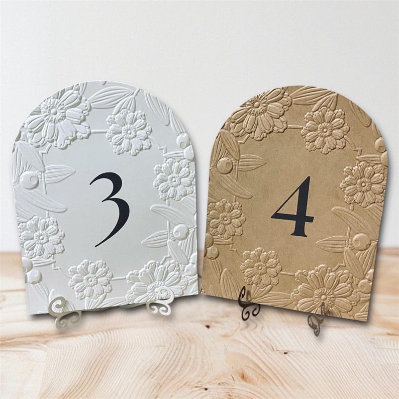May include: Two decorative table numbers, featuring a floral design. One is white with the number 3 in black, the other is tan with the number 4 in black. Both have a curved top and decorative metal stands.