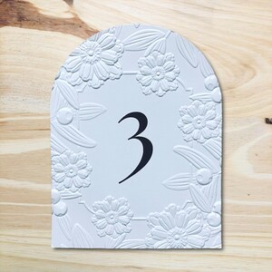 May include: White table number 3 with an arched top. The number is in black, centered on the card. The border is decorated with embossed flowers and leaves. The background is a light wood grain.