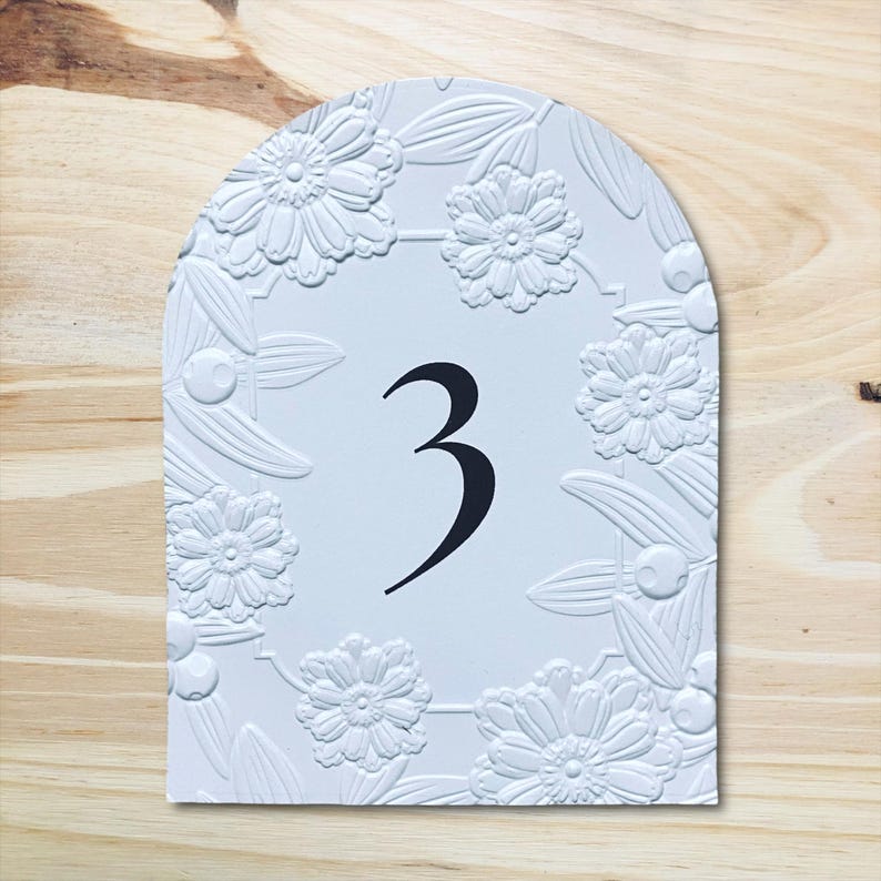 May include: White table number 3 with an arched top. The number is in black, centered on the card. The border is decorated with embossed flowers and leaves. The background is a light wood grain.