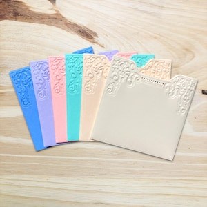 Peut inclure: Un ensemble de sept pochettes en papier rectangulaires dans diverses couleurs pastel, notamment bleu, lavande, rose, turquoise, pêche et crème. Chaque pochette présente un motif floral en relief sur les coins supérieurs et un bord perforé.