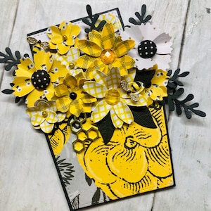 Könnte beinhalten: Eine gelbe und schwarze Karte mit einem Strauß gelber und schwarzer Papierblumen. Die Blumen sind in einer Vase mit einem Wabenmuster angeordnet. Die Karte hat einen schwarzen Rand und einen gelben und schwarzen gemusterten Hintergrund.