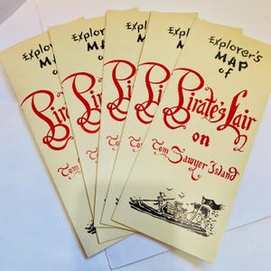 May include: A stack of cream-colored paper maps with red and black text. The maps read "Explorer's Map of Pirate's Lair on Tom Sawyer Island." A black illustration of a boat is at the bottom.