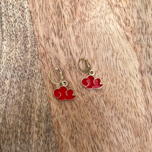 Puede incluir: Un par de pendientes de oro con dijes en forma de nube roja. Los pendientes están sobre una superficie de madera.