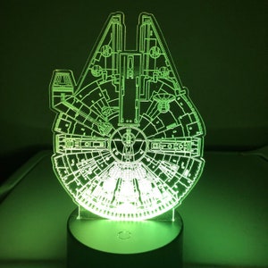 May include: A green-glowing, detailed acrylic night light shaped like the Millennium Falcon starship. The light sits on a black base, illuminating the ship's intricate design. The overall theme is science fiction.