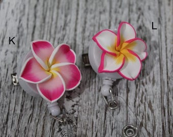 May include: Two white retractable badge holders with a flower design. One flower is pink and white, the other is white with a yellow center. Both flowers have a pink outline.