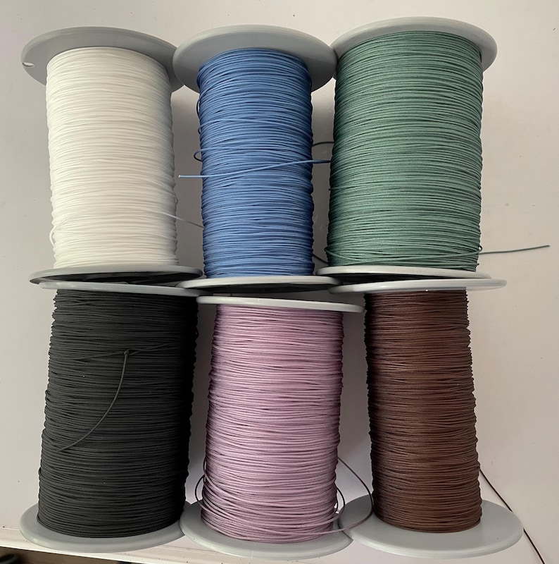 May include: Six spools of thread in different colours: white, blue, green, black, pink, and brown. The spools are all the same size and are arranged in two rows of three.
