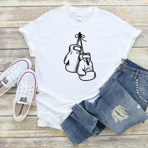 May include: White t-shirt with a black graphic of two boxing gloves hanging from a hook. The t-shirt is laid out on a wooden surface with a pair of white sneakers and blue jeans.