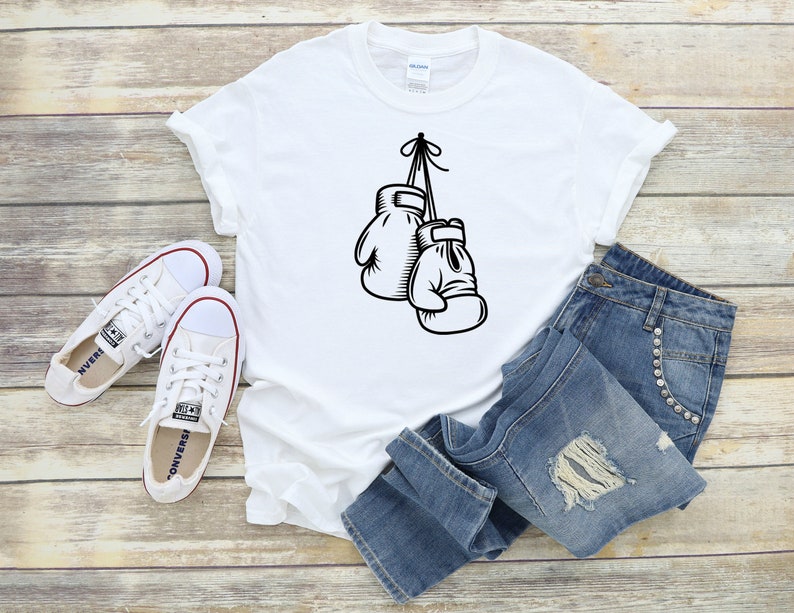 May include: White t-shirt with a black graphic of two boxing gloves hanging from a hook. The t-shirt is laid out on a wooden surface with a pair of white sneakers and blue jeans.