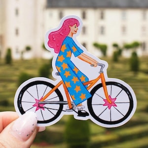 Peut inclure: Autocollant représentant une femme aux cheveux roses à vélo sur un vélo orange. Elle porte une robe bleue à motifs d'étoiles oranges. Un château et un jardin sont visibles en arrière-plan.