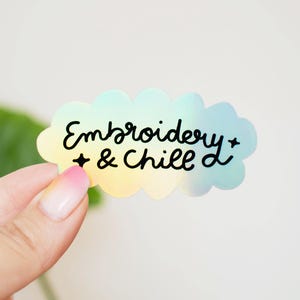May include: A cloud-shaped sticker with a pastel gradient background transitioning from yellow to blue. The sticker features the black text "Embroidery & Chill" written in a cursive script, accented by two small black four-pointed star symbols.