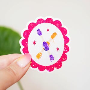 May include: A scalloped oval sticker with a bright pink holographic border featuring a white center. The design displays two crossed skeins of embroidery floss in purple and orange, accented by small red star shapes scattered around the center.