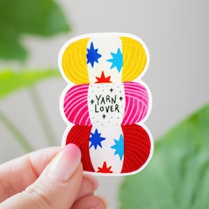 May include: A die-cut sticker featuring a stack of three yarn balls in yellow, pink, and red. A white paper band wraps around the center, printed with the text YARN LOVER, surrounded by small black stars and blue and red star-shaped icons.