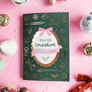 May include: A green notebook with gold illustrations and the text "Pause Créative Special Noël" is surrounded by Christmas decorations on a pink background. The decorations include white and gold baubles, acorn-shaped ornaments, and a velvet bow.