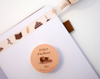 May include: A pink and brown washi tape roll with the text "lecture d'automne" and an illustration of a person sitting in a chair reading a book. The tape is 10 mm wide and 10 metres long.