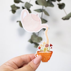 Op de afbeelding: Een witte theepot met een gespikkelde textuur giet thee in een bruine theekop met een gespikkelde textuur. De theekop is gevuld met bloemen en een paar aren tarwe.