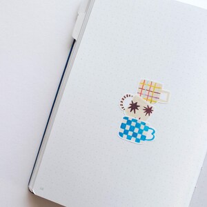 May include: A white notebook with a dotted pattern and a blue spine. Three colorful stickers of mugs are placed on the page. The mugs are stacked on top of each other. The top mug is yellow and red plaid, the middle mug is brown with white stars, and the bottom mug is blue and white checkered.