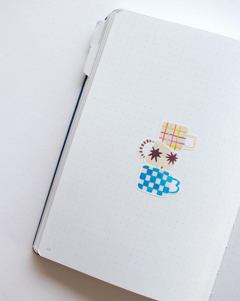 May include: A white notebook with a dotted pattern and a blue spine. Three colorful stickers of mugs are placed on the page. The mugs are stacked on top of each other. The top mug is yellow and red plaid, the middle mug is brown with white stars, and the bottom mug is blue and white checkered.