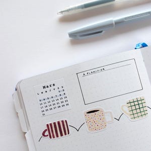 May include: A white planner with a calendar for March, a section labeled "A PLANIFIER", and a row of colorful coffee cup stickers.