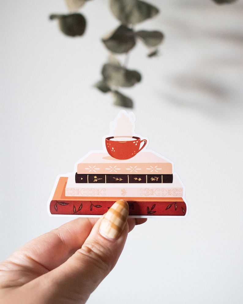 May include: A sticker of a stack of three books with a red mug of coffee on top. The books have a pink, black, and red cover with a floral design.