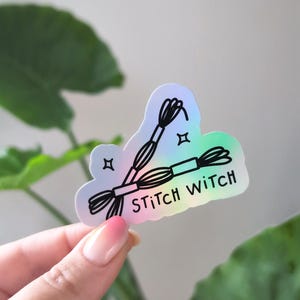 May include: A die-cut sticker with a holographic finish featuring a black line art illustration of two skeins of embroidery floss and the text STiTcH WiTcH. The design includes small four-pointed star accents on a gradient background.