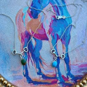 May include: Silver necklaces with a horse bit pendant, each featuring a gemstone. One necklace has a green stone, and another has a turquoise stone. The necklaces are displayed on a decorative tray with a colorful horse painting.