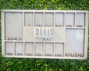 May include: A light-coloured wooden photo display board for school years, with spaces for photos from Pre-K to Senior year. The name "ELLIE" is in white, with "mac" below it. The board has a natural wood grain and is held together with metal screws.