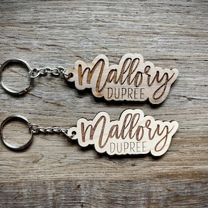May include: Two wooden keychains with the name "Mallory Dupree" engraved in a stylish font. The keychains are connected to a silver chain and ring. The keychains are made of light-colored wood and are set against a wooden background.