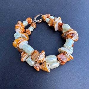 May include: A bracelet with a mix of orange and white shell-like beads and pale green oval beads. The bracelet is connected with a silver-coloured clasp. The beads have a natural, organic appearance.