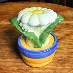 Puede incluir: Una flor de cerámica blanca con hojas verdes y un centro amarillo, sentada sobre una base de cerámica azul y amarilla. La flor tiene forma de margarita y tiene un aspecto realista.