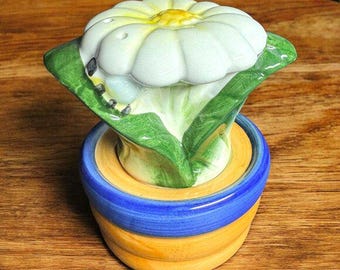 May include: A white ceramic flower with green leaves and a yellow center, sitting on a blue and yellow ceramic base. The flower is shaped like a daisy and has a realistic appearance.